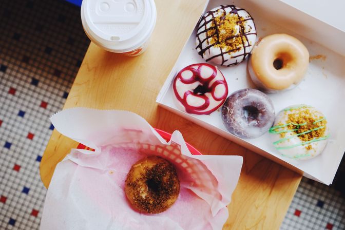 Top-down view of assorted colorful gourmet donuts in a white box and one donut in pink paper beside a takeaway coffee cup on a light wooden table above a tiled floor