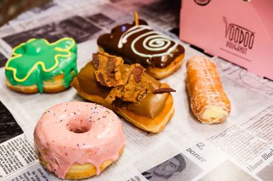 Assorted colorful gourmet donuts—pink sprinkled ring, green iced novelty, maple bar topped with crispy bacon, chocolate swirl bar, and sugar-coated cream-filled stick—arranged on newspaper-lined parchment.