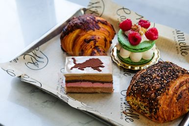Artisan bakery tray of pastries on parchment: chocolate-swirl croissant, seeded multigrain croissant, layered mousse cake slice with cocoa lion stencil, and a green raspberry macaron tower.