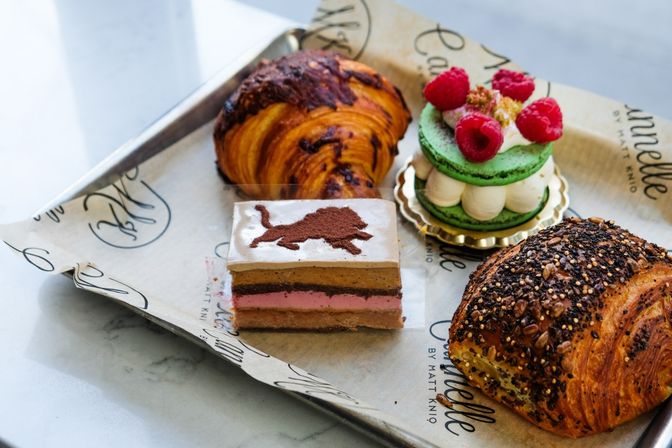 Artisan bakery tray of pastries on parchment: chocolate-swirl croissant, seeded multigrain croissant, layered mousse cake slice with cocoa lion stencil, and a green raspberry macaron tower.