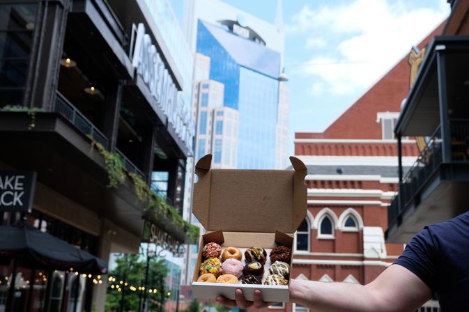 Outstretched arm holding an open takeout box of colorful assorted mini donuts against a downtown street with a glass skyscraper and red-brick building in the background.