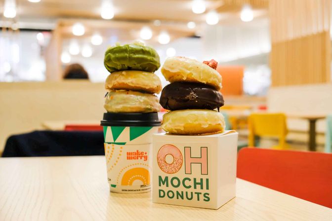 Colorful mochi donuts stacked beside a patterned takeaway coffee cup on a light wood table in a bright indoor café with blurred bokeh lights and colorful chairs.