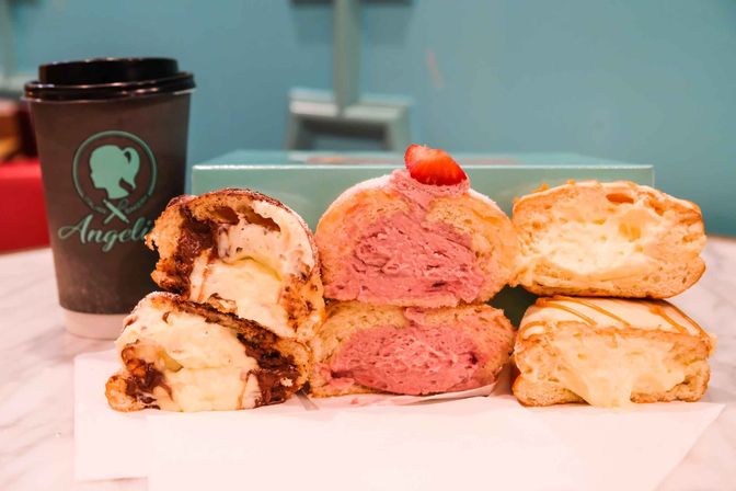 Close-up of cut cream puffs: chocolate-swirl vanilla cream, bright pink strawberry cream topped with a strawberry slice, and caramel-drizzled custard, with a teal takeaway coffee cup and pastel box in the background.