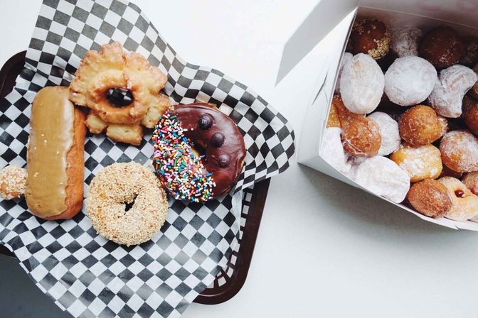 Top-down view of assorted donuts — chocolate-sprinkled, sesame-covered, glazed long john and an apple fritter — on a black-and-white checkered paper-lined tray next to an open box of powdered donut holes on a white table.