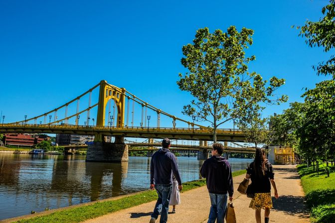 Sunny riverfront path with three people walking past a bright yellow suspension bridge over a calm river, green trees and clear blue sky.