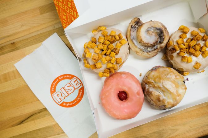 White box of assorted breakfast pastries on a wooden table — glazed and pink jam-filled donuts, a cinnamon roll, and two cereal-topped donuts.