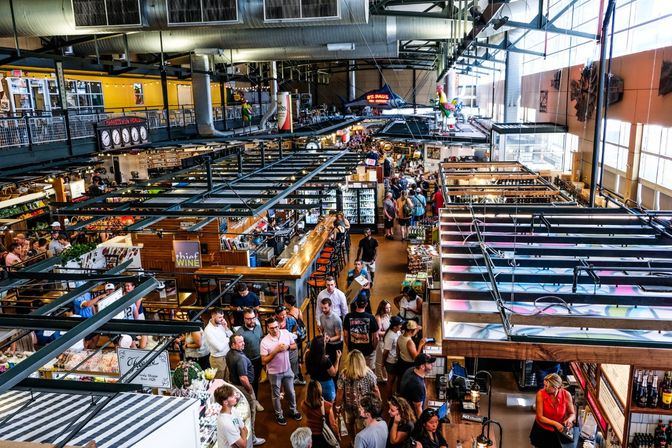 Bustling urban indoor market in a loft-style hall — crowded food stalls and vendor booths, mezzanine seating, wine bar, exposed metal beams and large windows.