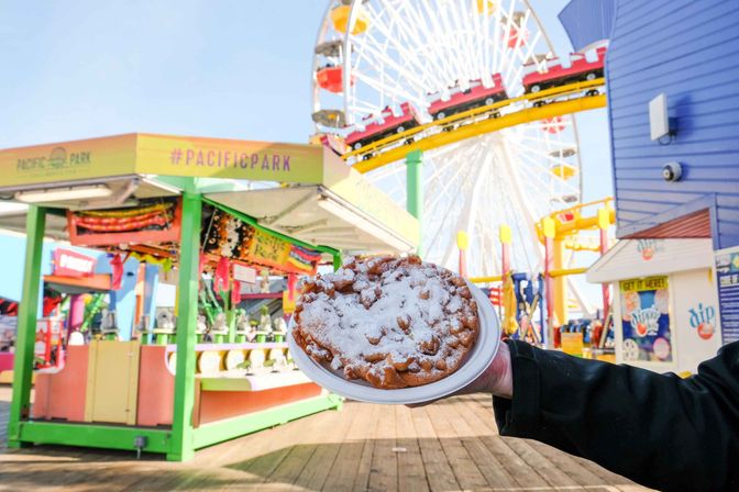 Hand holding a powdered-sugar funnel cake on a paper plate at a colorful seaside boardwalk amusement park with a Ferris wheel and roller coaster in the sunny background