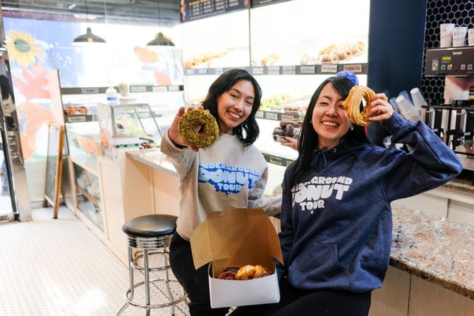 Two smiling people in hoodies holding donuts and a box of assorted donuts while sitting at a bright donut shop counter with pastry display cases and a coffee station