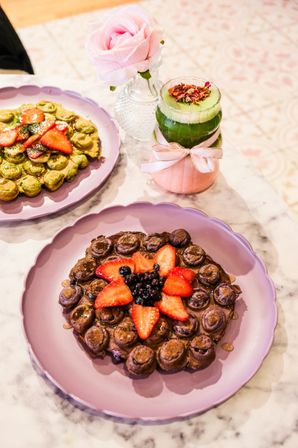 Pretty cafe brunch scene: pink plates with bubble waffles topped with sliced strawberries and berries on a marble table, paired with a layered matcha drink in a ribbon-tied jar and a pink rose