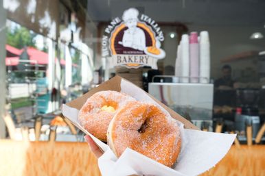 Hand holding two sugar-coated filled donuts in a paper tray outside a bakery window with stacked coffee cups and pastry counter visible inside
