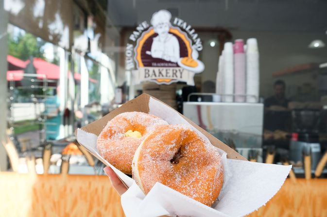 Hand holding two sugar-coated filled donuts in a paper tray outside a bakery window with stacked coffee cups and pastry counter visible inside