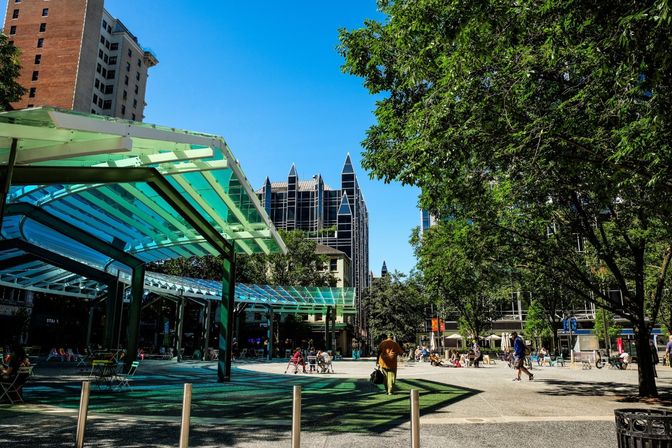 Sunny downtown plaza with turquoise glass canopy, scattered chairs and tables, leafy trees and people strolling beneath pointed modern skyscrapers