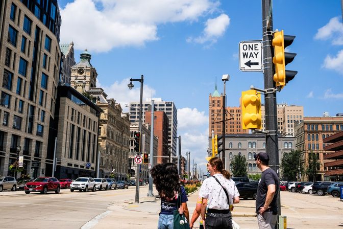 Downtown urban intersection with pedestrians waiting at a crosswalk, yellow traffic lights and a one-way sign, lined with a historic clock tower and modern office buildings under a bright blue sky.