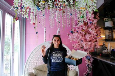 Smiling woman holding a donut and an open box of assorted donuts inside a pink, floral Instagrammable cafe with hanging white wisteria, decorative butterflies and a white wicker swing chair.