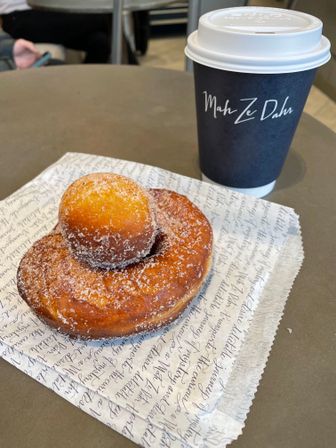 Sugar-dusted yeast donut topped with a doughnut hole next to a takeaway coffee cup with a script logo, served on patterned wax paper at a café table.