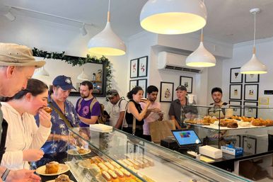 Customers queue at a bright neighborhood bakery counter, choosing pastries from a glass display case stocked with éclairs, croissants and tarts under white pendant lights and framed art.
