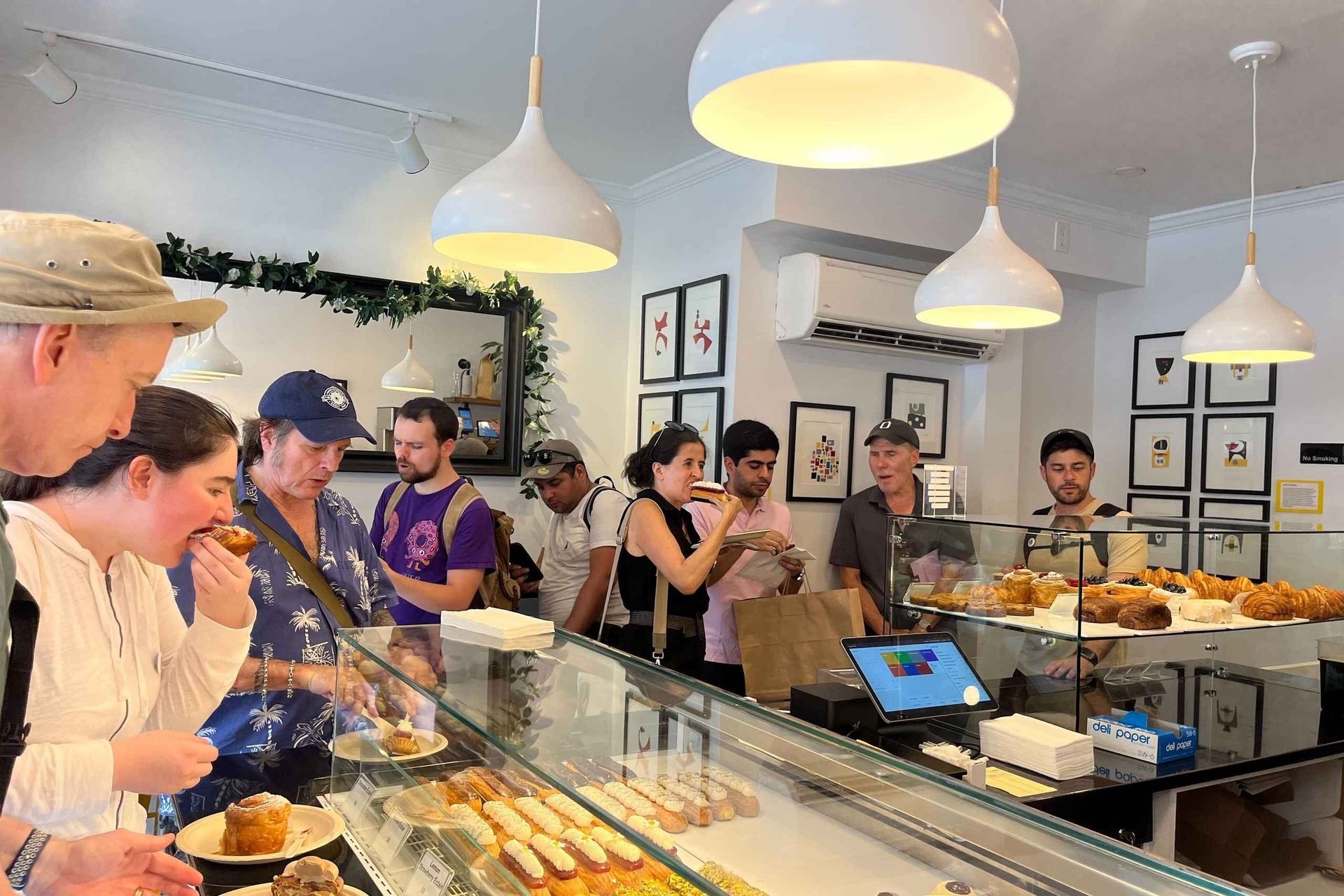 Customers queue at a bright neighborhood bakery counter, choosing pastries from a glass display case stocked with éclairs, croissants and tarts under white pendant lights and framed art.