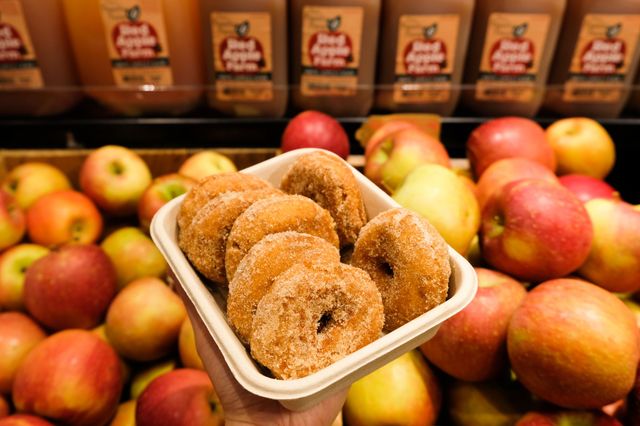 Tray of cinnamon-sugar apple cider donuts held above a pile of fresh apples and apple cider bottles in a grocery produce display, autumn snack