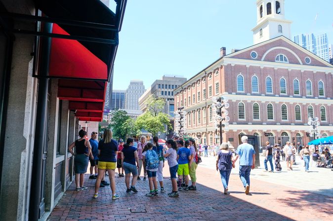 Sunny summer scene in downtown Boston: tourists gather on a brick-paved pedestrian square under red awnings, with a historic red-brick market hall topped by a white cupola in the background.