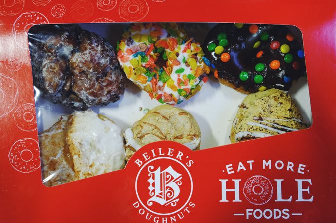 Red takeout box with six assorted colorful doughnuts visible through a window — glazed, cereal-topped, chocolate with candy pieces, and drizzled varieties for a fun breakfast treat.