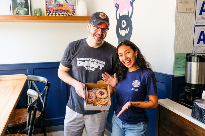 Two smiling staff in a bright neighborhood donut shop holding an open box of assorted gourmet donuts (pink glazed and pastry) by blue wainscoting, playful wall mural and coffee station