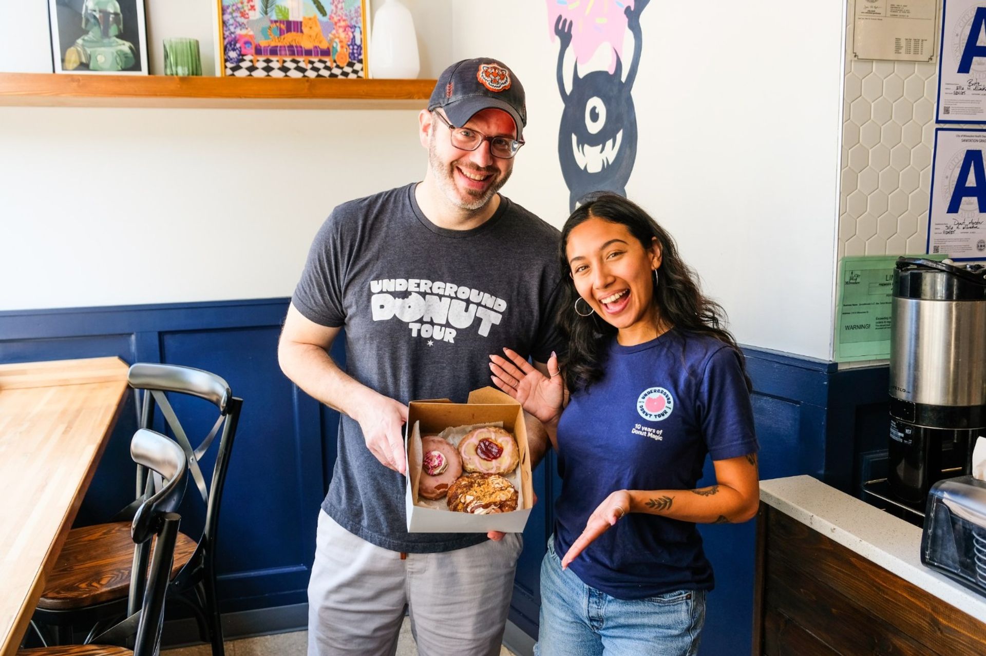 Two smiling staff in a bright neighborhood donut shop holding an open box of assorted gourmet donuts (pink glazed and pastry) by blue wainscoting, playful wall mural and coffee station