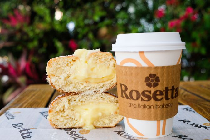 Custard-filled bakery bun stacked beside a takeaway coffee cup with a brown cardboard sleeve on a wooden outdoor cafe table, lush green and pink foliage blurred in the background.