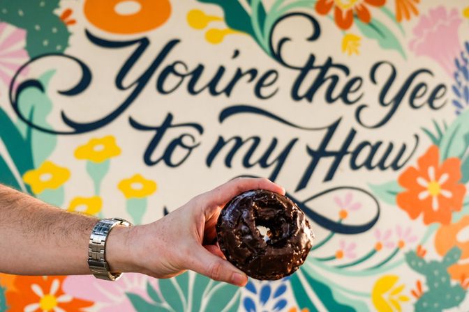 Hand holding a chocolate-glazed donut in front of a colorful floral mural with cursive text 'You're the Yee to my Haw'.