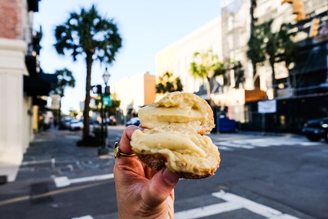 Hand holding a split cream-filled pastry in the foreground with a sunlit, palm-lined downtown street, blurred buildings and a crosswalk in the background.