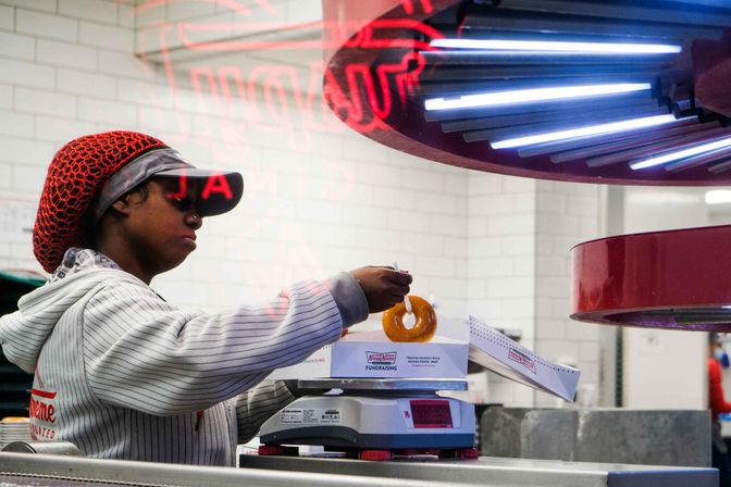 Bakery worker in a red hairnet and visor places a glazed donut into a box on a scale at a donut shop counter under blue neon lights