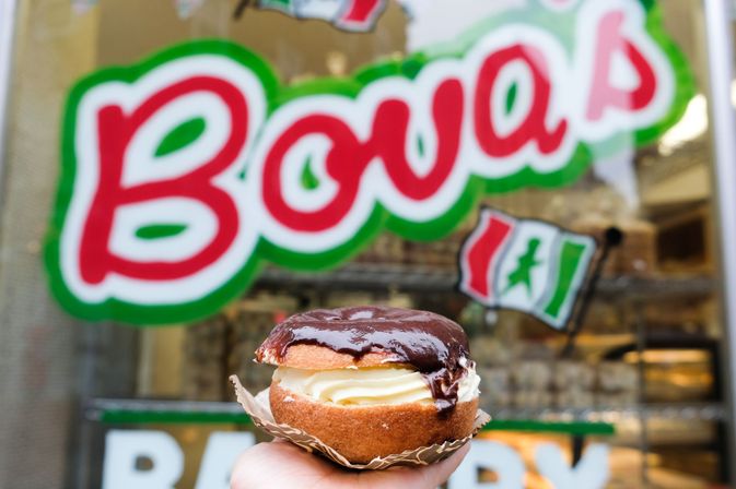 Decadent cream-filled, chocolate-glazed round pastry held by hand in front of a vibrant bakery window with green-and-red signage