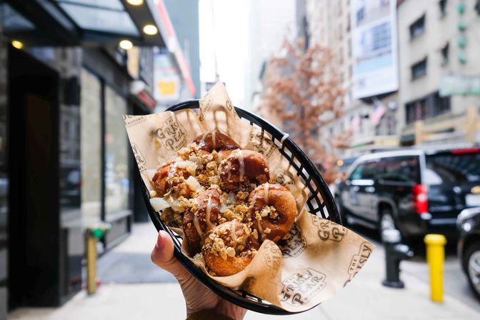 Hand holding a basket of warm glazed mini doughnuts topped with caramel drizzle and crunchy crumble, blurred urban street and parked cars in the background