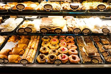 Bright glass bakery display case with rows of assorted pastries — croissants, cream-filled rolls, cronuts, glazed donuts and pecan bars.