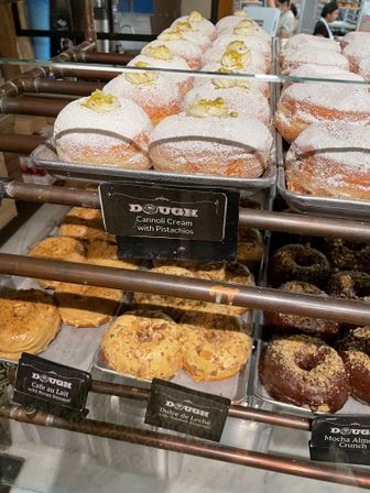 Glass bakery display case of assorted gourmet donuts — powdered cannoli-cream donuts topped with chopped pistachios on the top tray and caramel-almond, dulce de leche and chocolate-crunch varieties on lower trays.