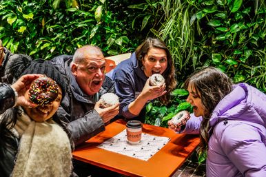 Four people at an outdoor cafe table with a living green wall, laughing and biting into colorful frosted donuts beside a takeaway coffee cup.