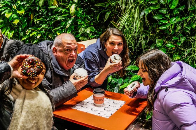 Four people at an outdoor cafe table with a living green wall, laughing and biting into colorful frosted donuts beside a takeaway coffee cup.