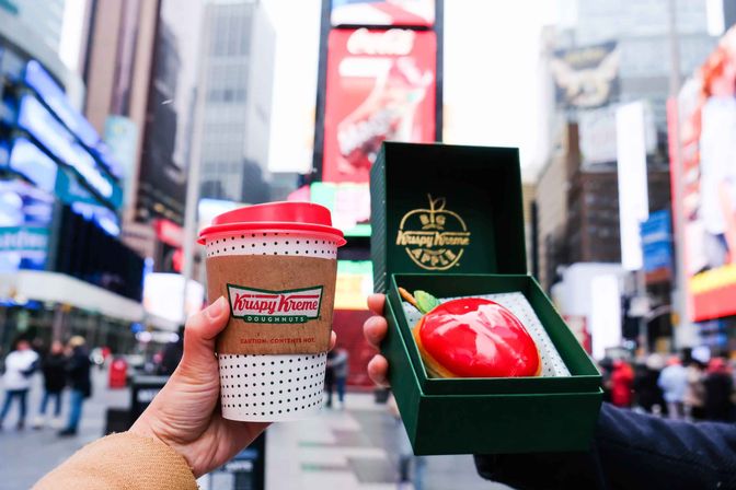 Takeaway coffee cup with red lid and green pastry box holding a glossy red apple-shaped doughnut, held up in Times Square, New York City with bright billboards and blurred crowds