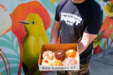 Person holding an open box of assorted gourmet donuts on a sunny sidewalk in front of a vibrant tropical bird street mural.