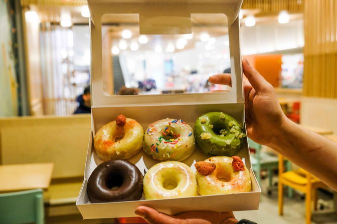 Open white box of six assorted gourmet donuts — chocolate-glazed, vanilla-glazed with rainbow sprinkles, matcha with crumble, and fruit-topped — held by hands in a bright modern café