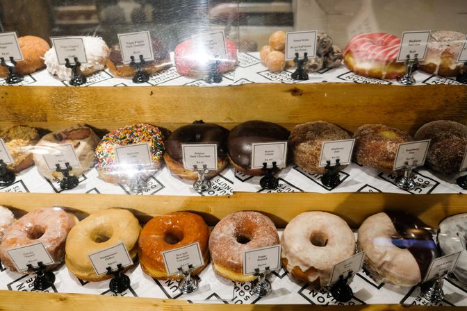 Colorful artisan donuts arranged in a wooden glass bakery display case — assorted glazed, sugar‑coated, chocolate‑topped, sprinkle and filled varieties with small menu labels.