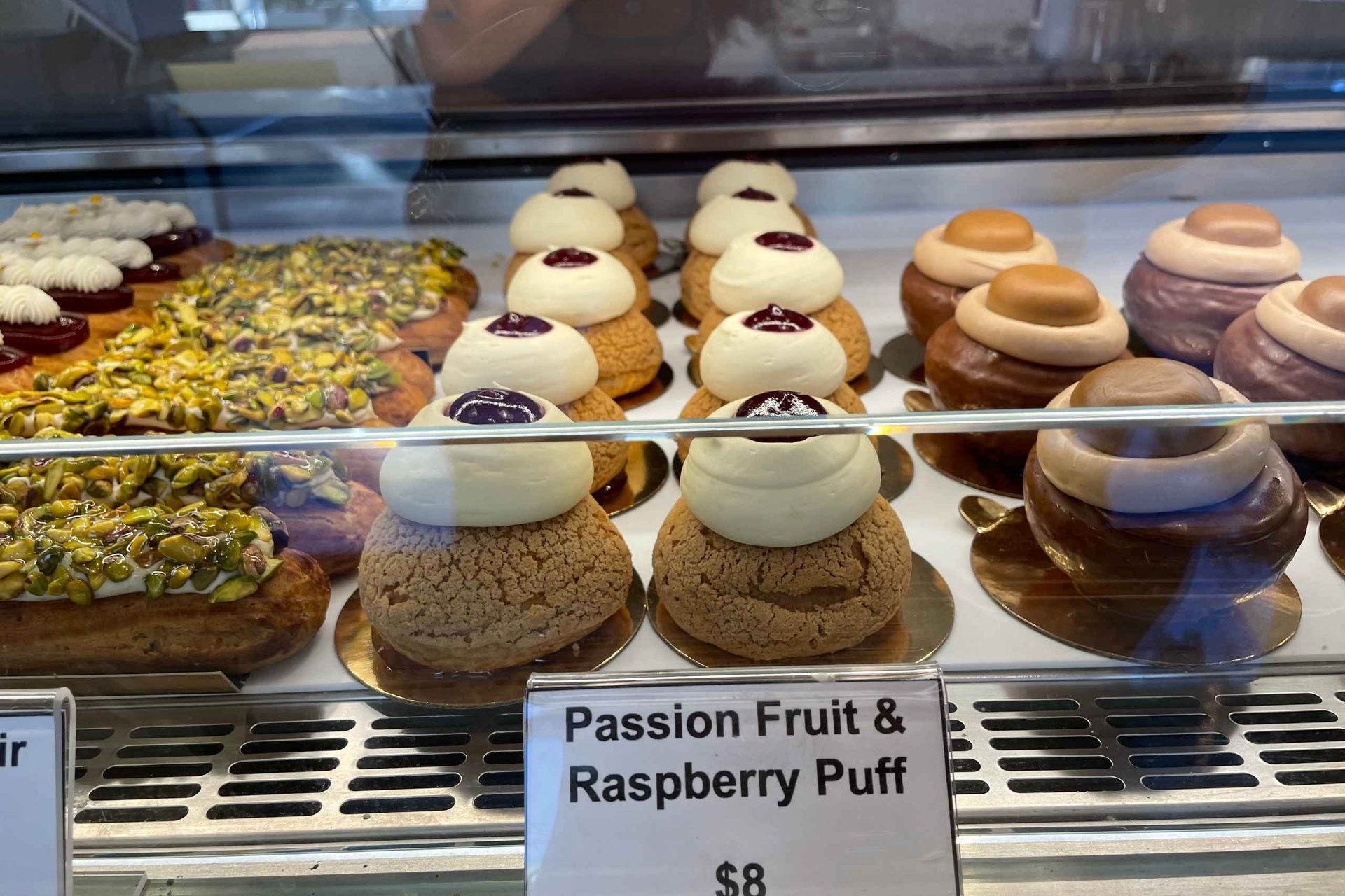 Glass bakery display case filled with rows of passion fruit and raspberry cream puffs topped with white cream and jam, pistachio‑topped éclairs and chocolate‑glazed pastries; sign reads $8.