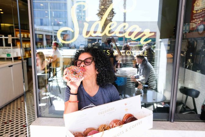 Person biting a large sprinkle-covered donut outside a city donut shop window, holding an open box of assorted donuts with café interior, patrons, and street reflections behind.