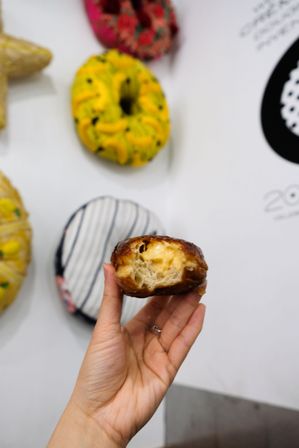 Hand holding a bitten cream-filled glazed doughnut with gooey custard, blurred colorful doughnut wall display in a modern bakery