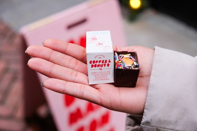 Close-up of an outstretched hand holding a tiny white box labeled “Coffee & Donuts” and a dark chocolate square topped with rainbow sprinkles and a mini donut, pink blurred background.