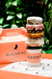Stack of four colorful doughnuts — white glazed with sprinkles, pink crushed topping, chocolate with sprinkles, coconut — balanced on a takeaway coffee cup beside a coral doughnut box against green foliage.