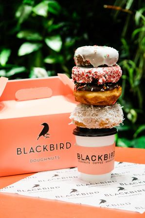 Stack of four colorful doughnuts — white glazed with sprinkles, pink crushed topping, chocolate with sprinkles, coconut — balanced on a takeaway coffee cup beside a coral doughnut box against green foliage.