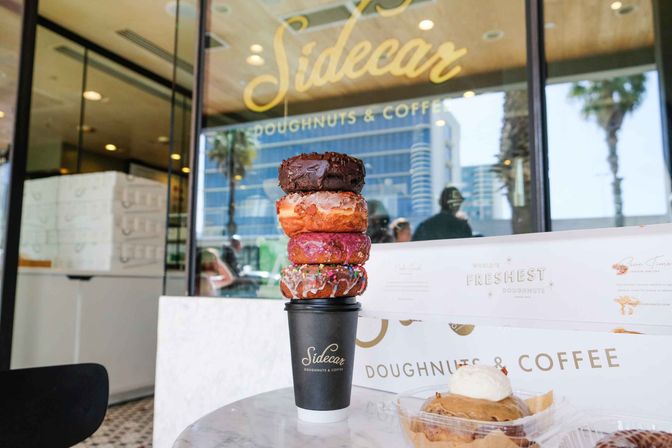 Four colorful doughnuts stacked on a black takeaway coffee cup on a marble table outside a beachside doughnut cafe, glass storefront reflecting palm trees and city buildings.