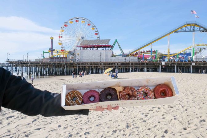 Box of assorted colorful donuts held on a sunny sandy beach with the Santa Monica Pier ferris wheel and roller coaster in the background