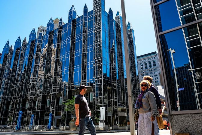 Sunlit downtown street with pedestrians in front of a blue mirrored glass office tower topped by pointed spires, reflective façades showing neighboring buildings and outdoor café seating with blue umbrellas